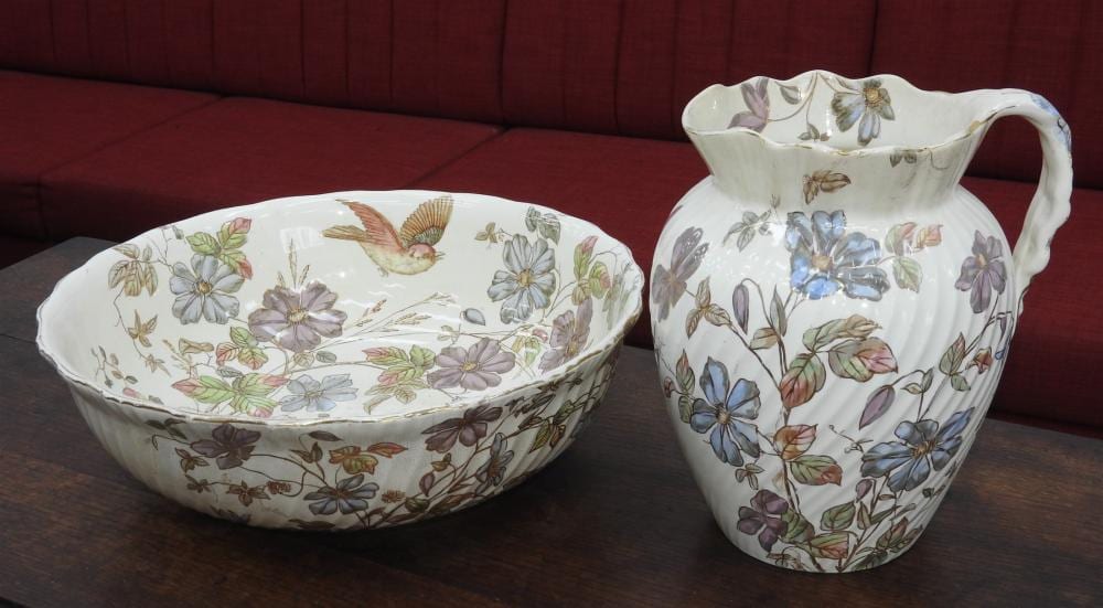 Porcelain wash basin and pitcher with painted floral decoration on a table in front of a red upholstered couch.
