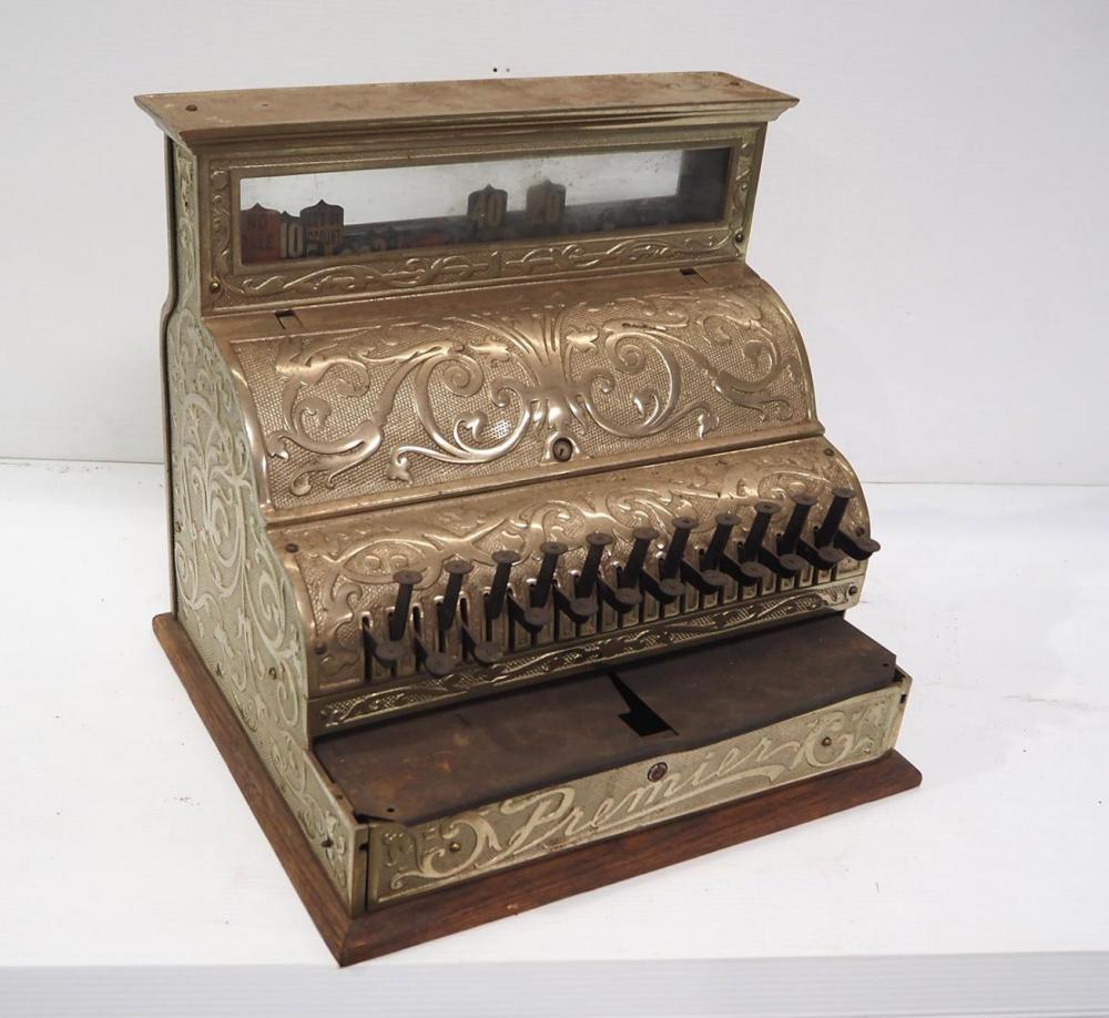 Brass cash register with ornate embossed panels, open top window, and rows of black numbered keys.
