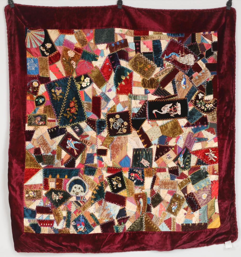 Square quilt top made of colorful fabric scraps and embroidered patches arranged in an irregular patchwork design, shown on a red backing.