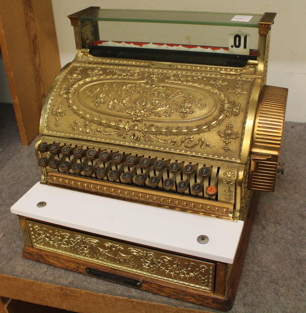 Vintage gold-colored cash register with ornate embossed front and a row of colorful number keys, shown on a countertop with drawer below.