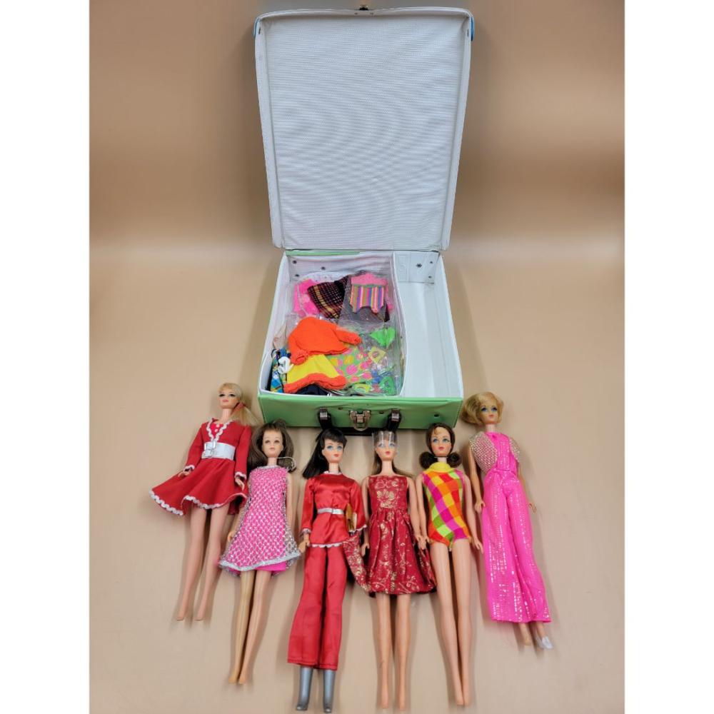 Vintage fashion dolls in colorful dresses displayed in front of an open green case with fabric pieces and accessories inside.