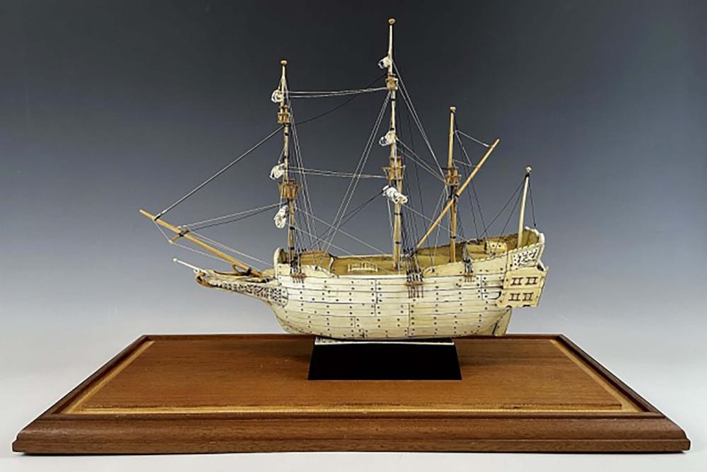 Antique-style miniature sailing ship model on a wooden display base with rigging and multiple masts against a dark background.