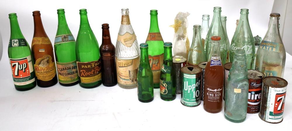 Collection of vintage soda bottles and glass drink bottles, including green and brown 7up bottles and a large clear bottle, arranged on a white surface.