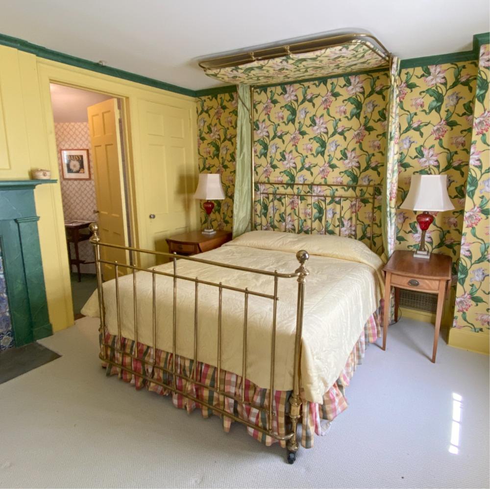 Antique brass canopy bed with floral fabric panels and a white bedspread in a bedroom corner.