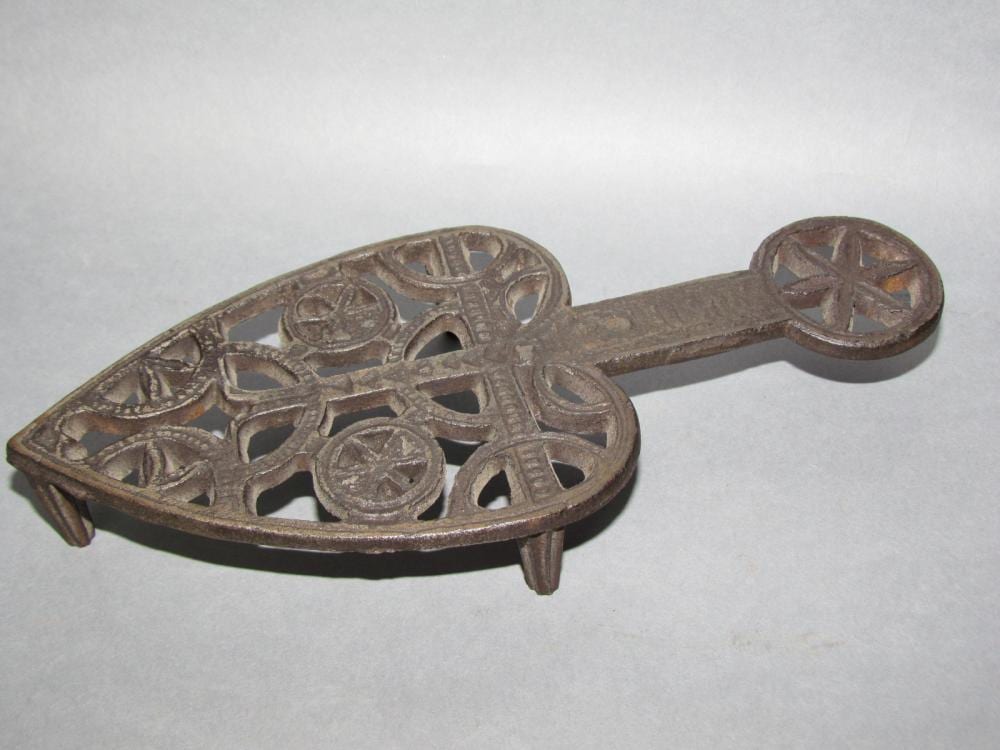 Cast iron trivet with circular star and geometric cutouts, shown from the side with a handle-like loop at one end.