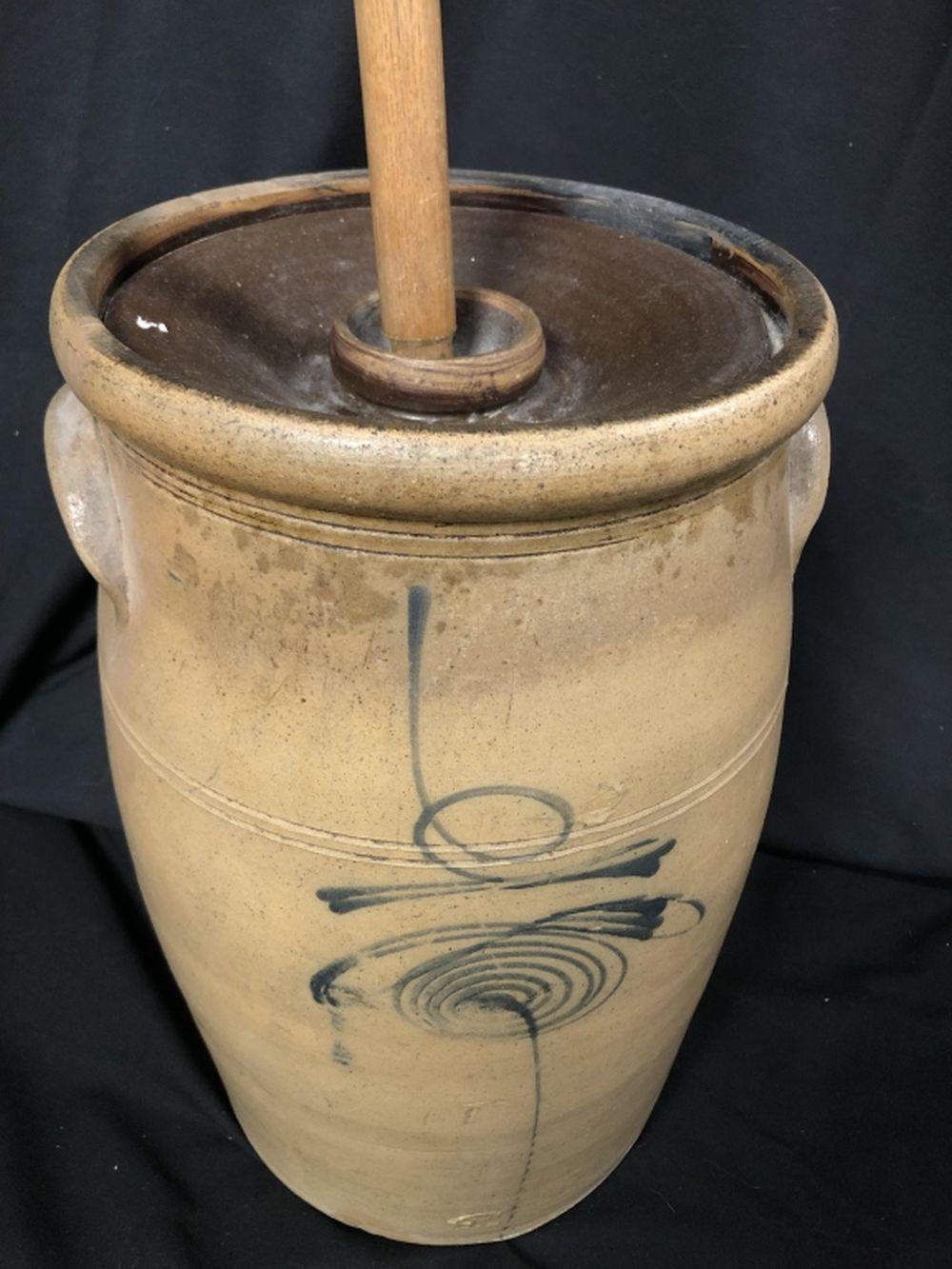 Stoneware crock-style butter churn with a wooden plunger handle and cobalt blue decorative designs against a dark background.