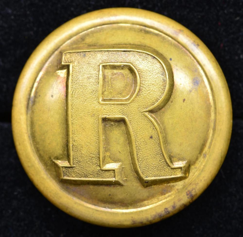 Close-up of a round brass-colored button with an embossed letter “R” design in the center.