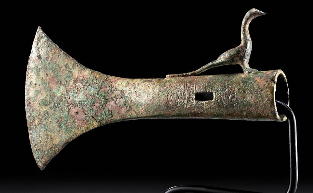 Ancient bronze axe head with patina, incised decoration near the blade and a curved looped socket.