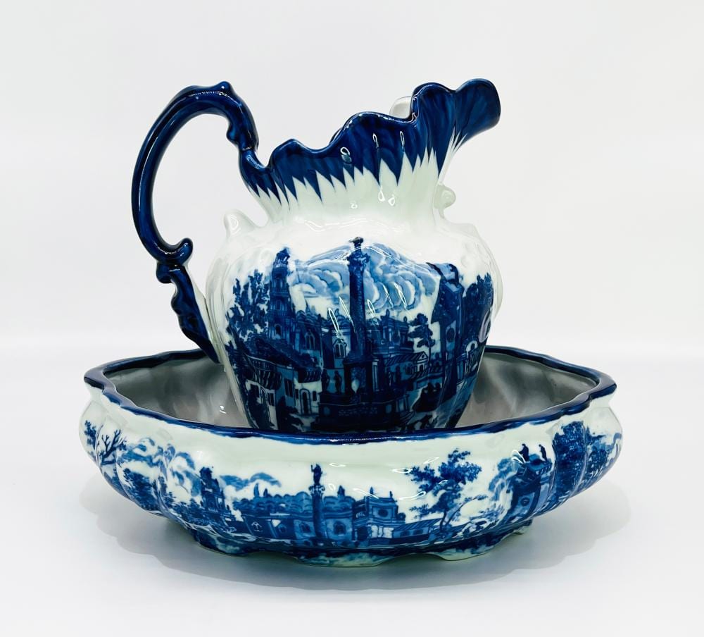 Blue and white ceramic pitcher with handle and matching wash basin bowl, decorated with a cityscape scene, on a white background.
