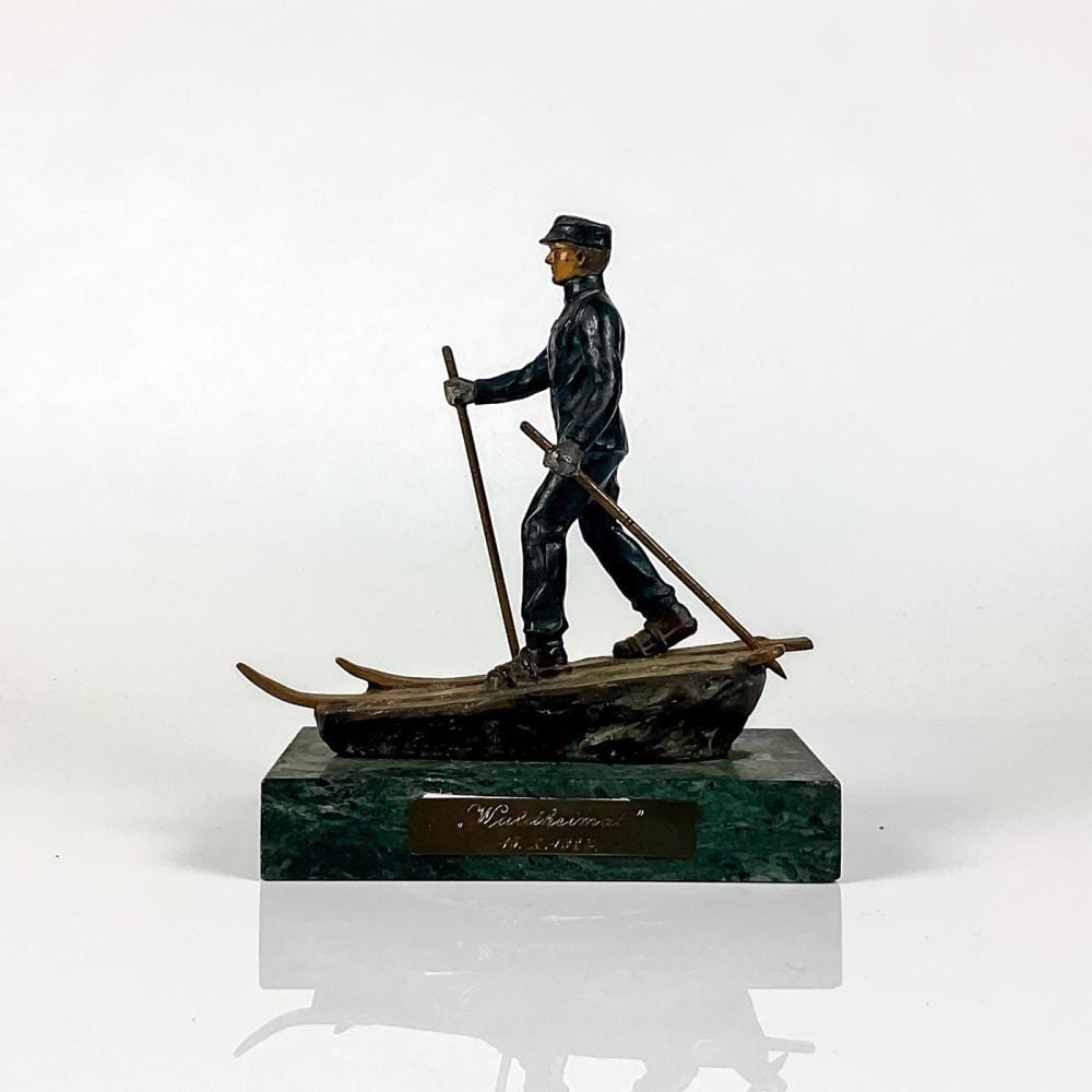 Bronze sculpture of a skier gliding forward while holding ski poles, mounted on a green stone base.