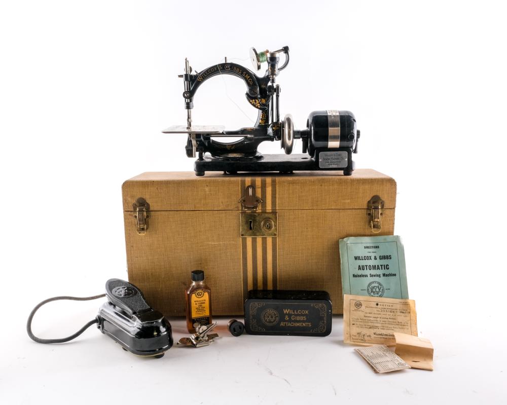 Front view of a vintage black sewing machine mounted on a tan fabric-covered case, with accessories and instruction papers on a white background.