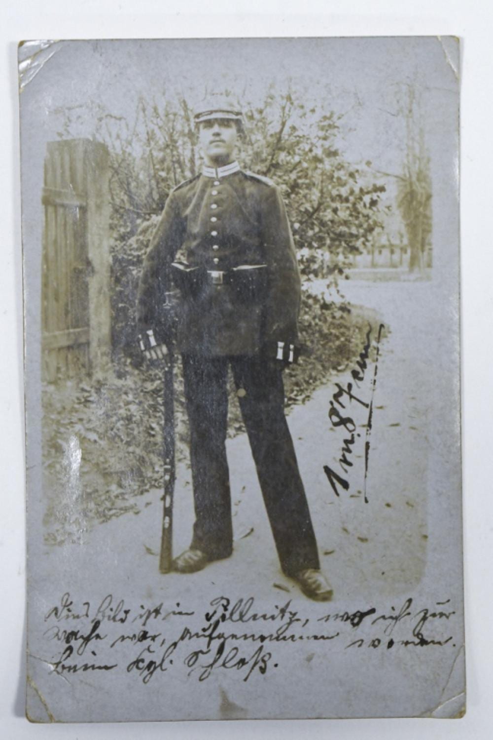 Black-and-white real photo postcard showing a seated man in a dark military-style uniform outdoors with plants behind him.