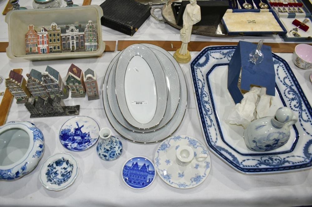 Tabletop display of blue-and-white ceramic serving dishes, platters, and lids, with a white figurine and decorative items in the background.