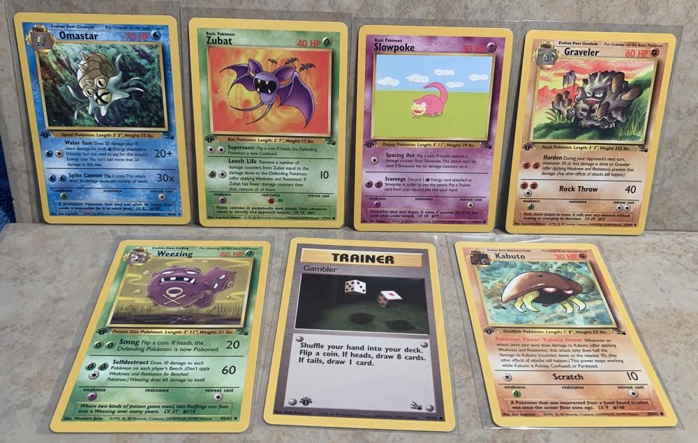 Photo of seven Pokémon trading cards on a gray tabletop, including cards labeled Omastar, Zubat, Slowpoke, Gravel­er, Weezing, Trainer, and Kabuto.