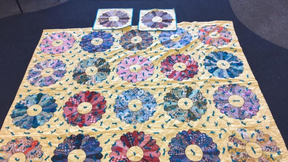 Vintage-style quilt top with colorful appliqué flowers and circular centers on a light cream background, shown flat on a dark surface.