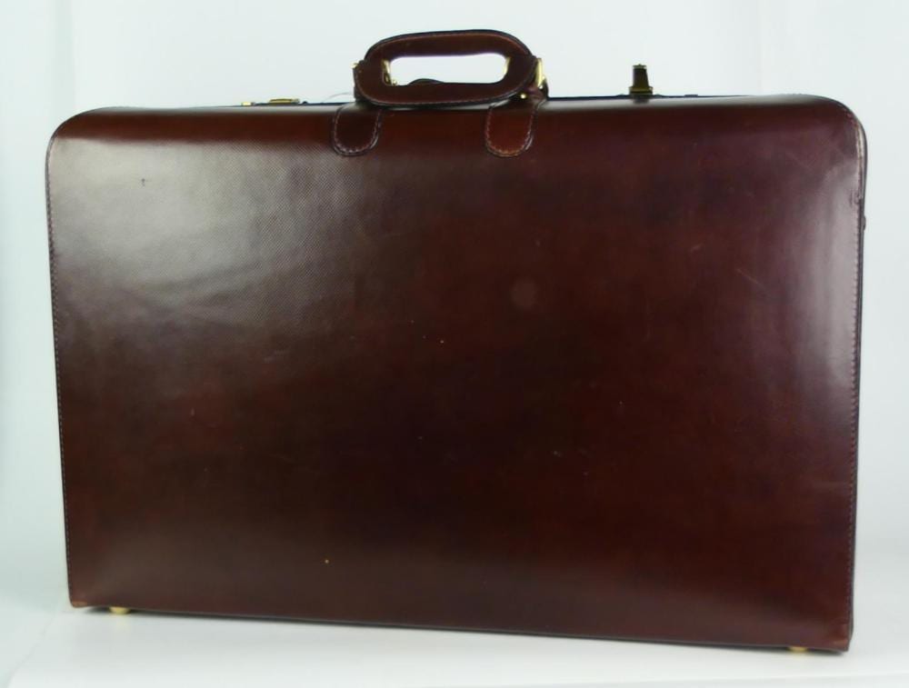 Brown leather suitcase with a top carry handle and side corners, shown against a white background.