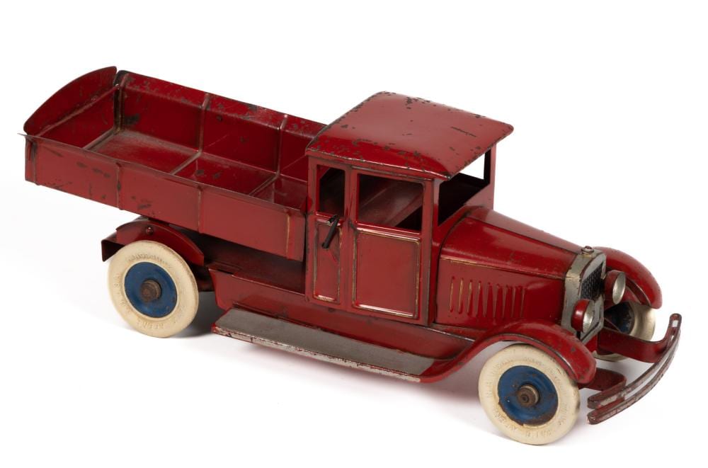 Red vintage wind-up dump truck toy with open cargo bed, detailed cab, and blue wheels on a white background.