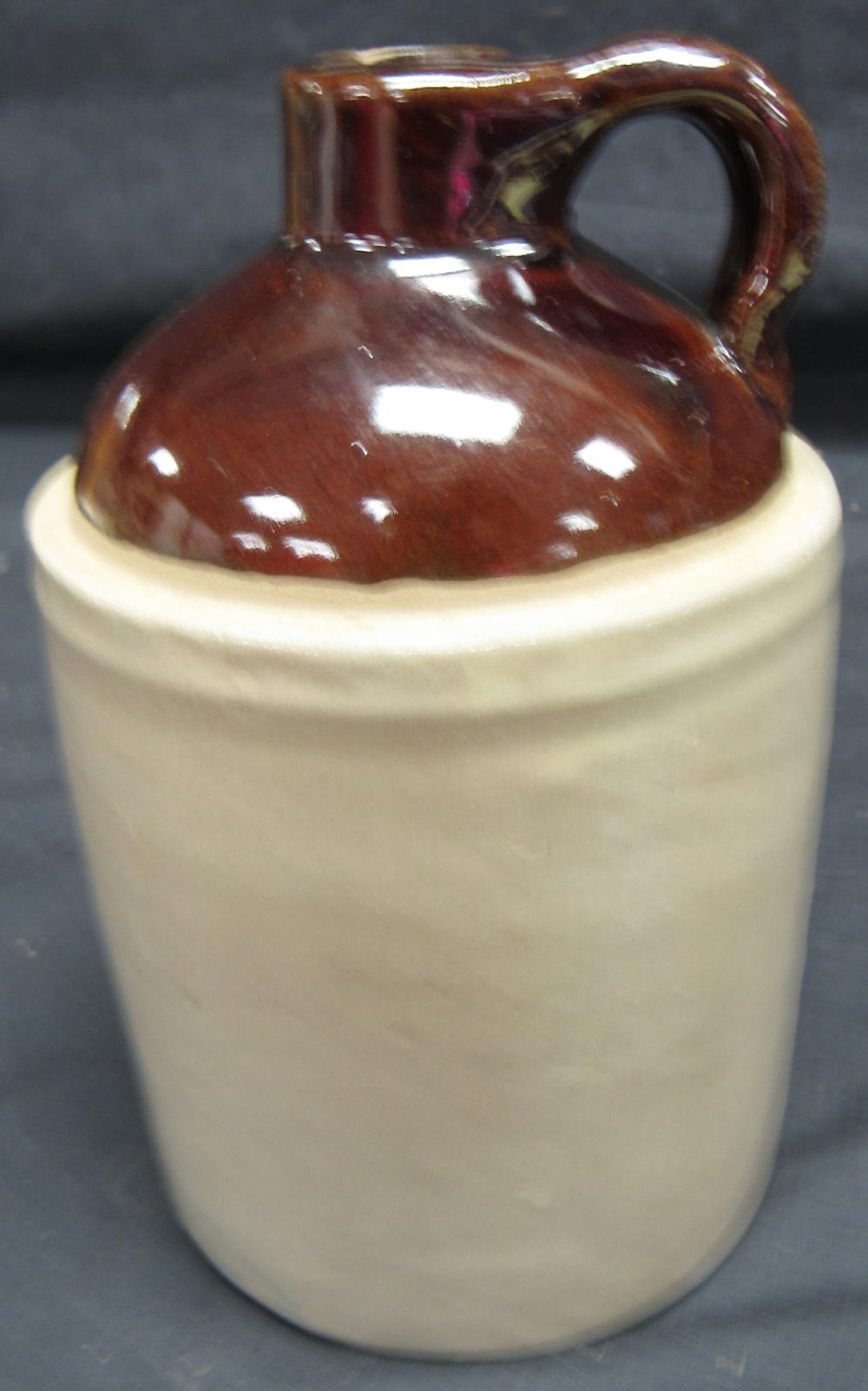 Brown two-tone stoneware whiskey jug with a handle and rounded body, shown on top of a cream-colored ceramic container.
