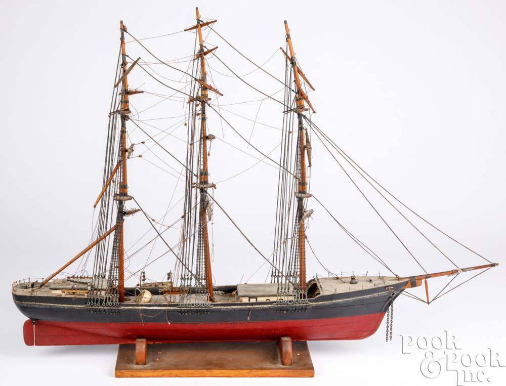 Wooden ship model with three masts, rigging lines, and a dark hull on a display stand against a white background.