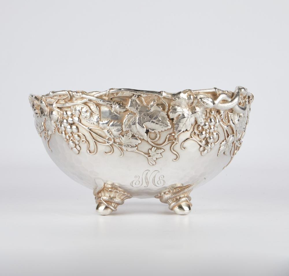 Silver tone punch bowl with grape and grapevine relief decoration and three curved feet, shown against a light background.