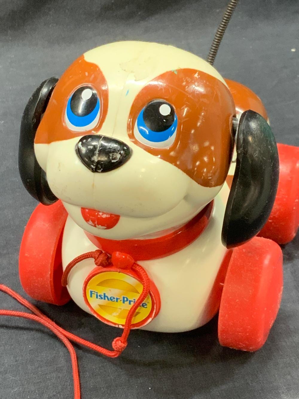 Red and white Fisher-Price dog pull toy with big blue eyes, black wheels, and an orange nose, shown on a gray surface.