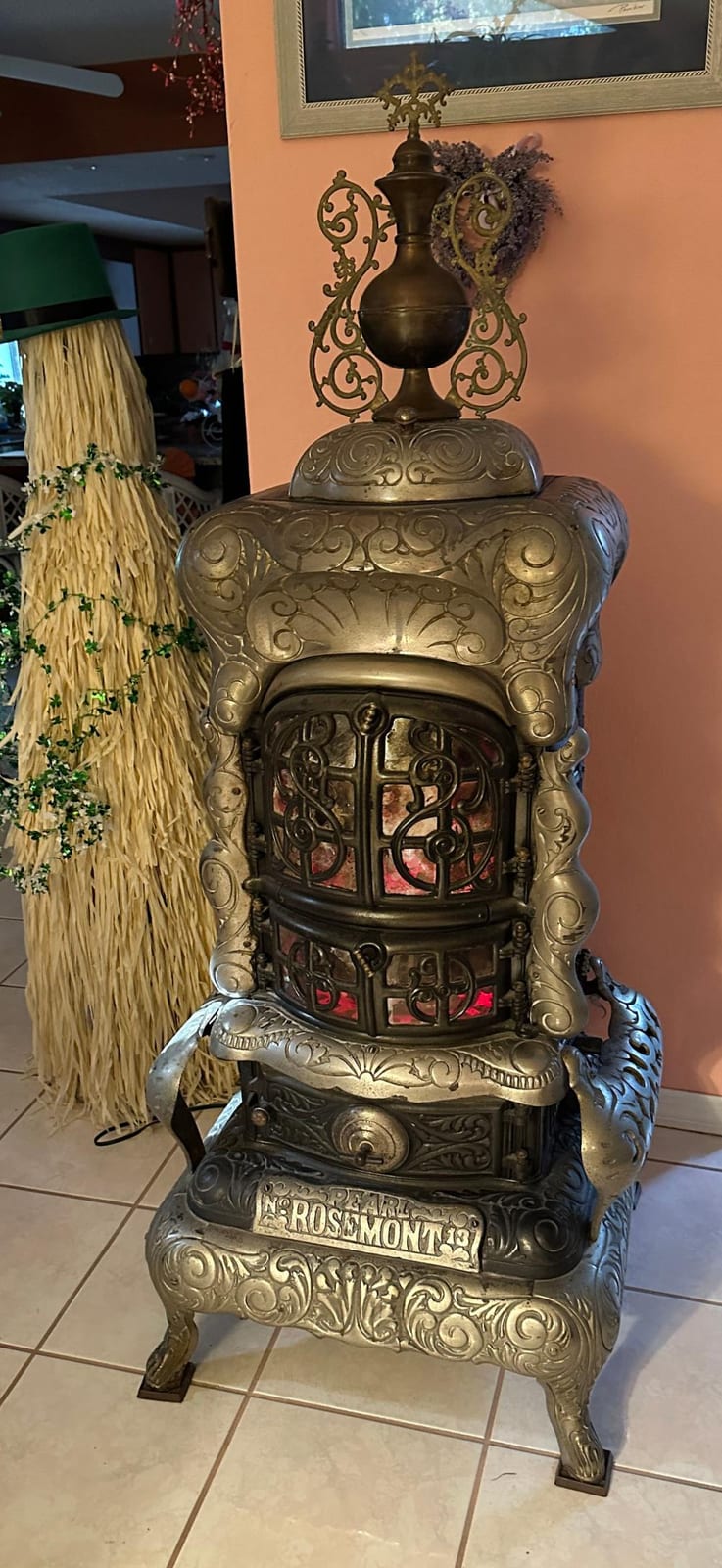 Brass decorative wood-burning stove with ornate scrollwork and raised top finial, shown indoors near a broom and wall art.