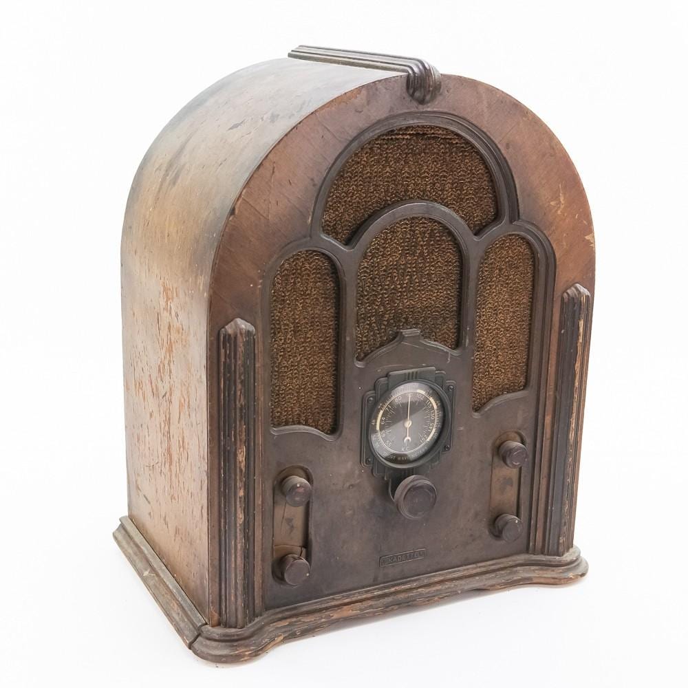 Front view of a vintage cathedral-style radio with a wood cabinet, brown speaker grille, round clock-like dial, and tuning controls.