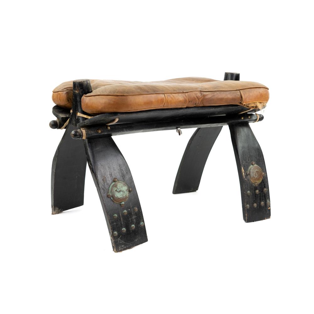 Vintage camel saddle-style upholstered ottoman stool with black wooden legs and bronze-colored studs, shown from the side on a white background.