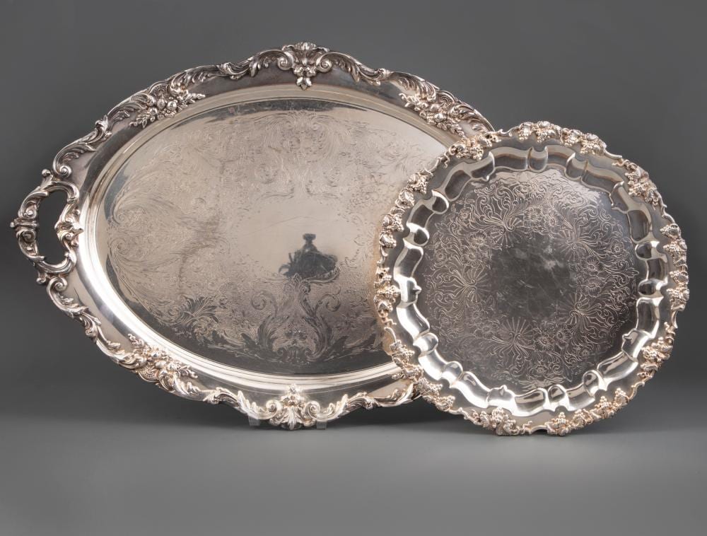 Two ornate silver-toned metal trays with raised scrolling borders and engraved floral patterns, shown on a dark background.