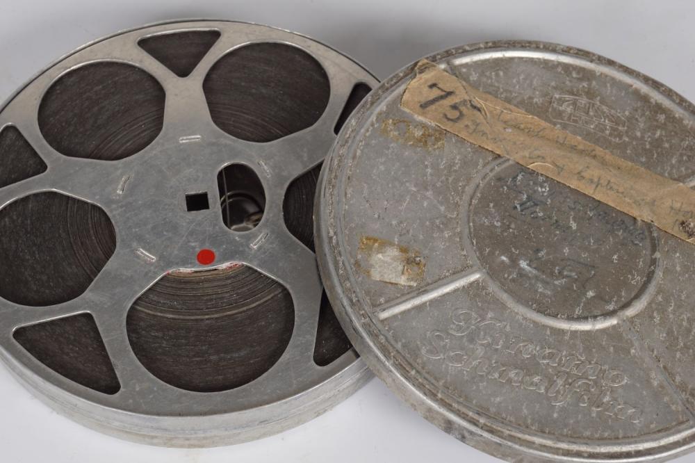 A gray 16mm film reel with dark film wound inside, shown beside its round metal can lid.