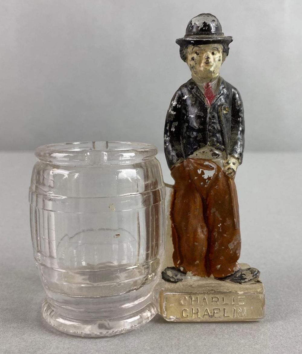 Clear glass toothpick holder shaped like a barrel next to a small painted figure of a man in a hat, on a light background.