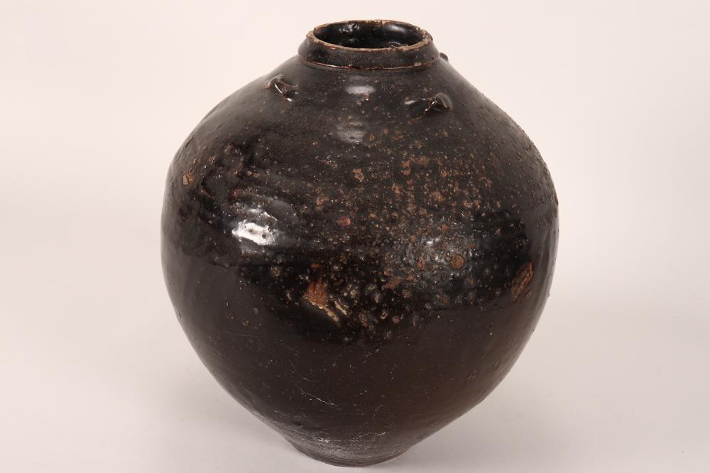 Small dark ceramic jar with a narrow neck and worn, speckled surface, shown against a plain light background.