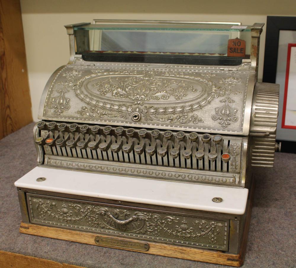 Vintage silver-toned cash register with ornate embossed front and keyboard-style number keys, shown on a tabletop