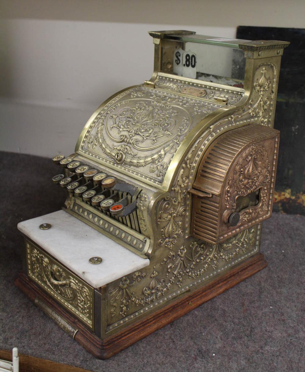 Antique brass cash register with ornate floral engraving, cash drawer open, and numbered keys visible in a row.