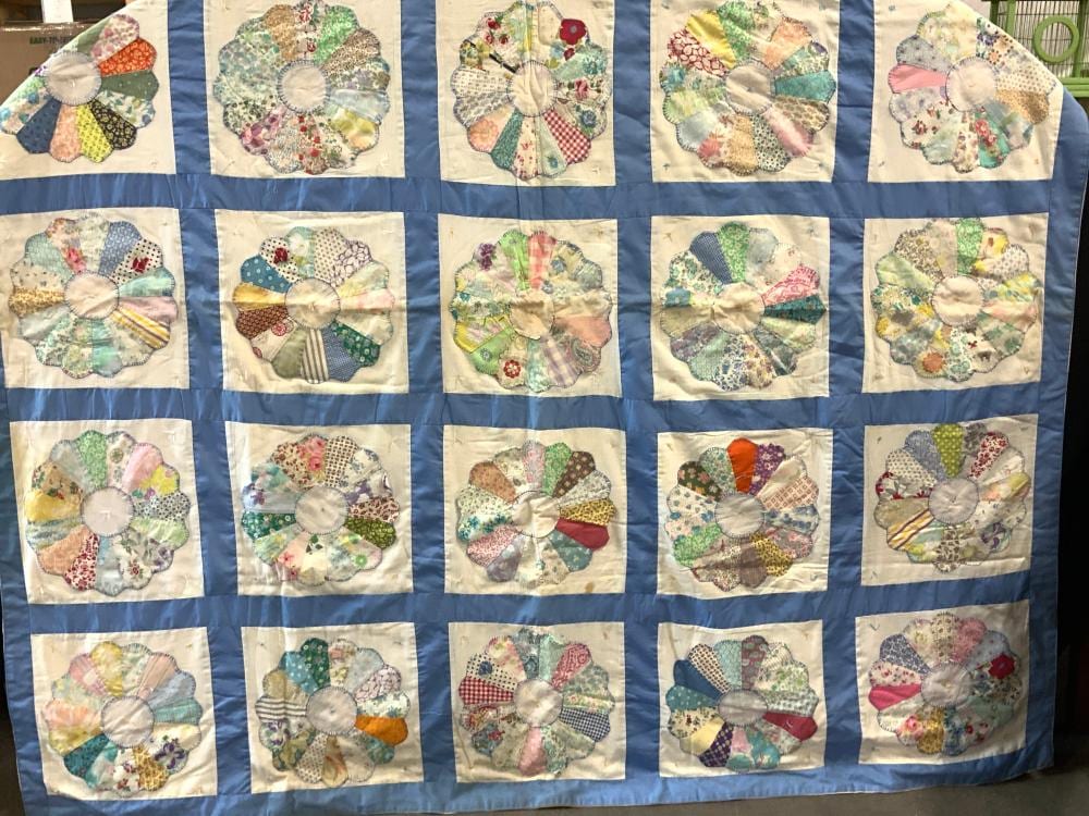 Quilt made of blue fabric squares with appliqué patchwork flower blocks, each featuring a fabric circle center and curved petal shapes in pastel colors.