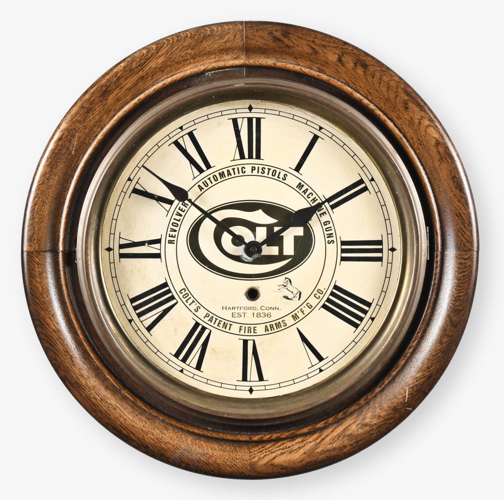 Round wall clock with a brown wood frame and a cream clock face featuring black Roman numerals and an “AUTOMATIC PISTOLS” advertising graphic.