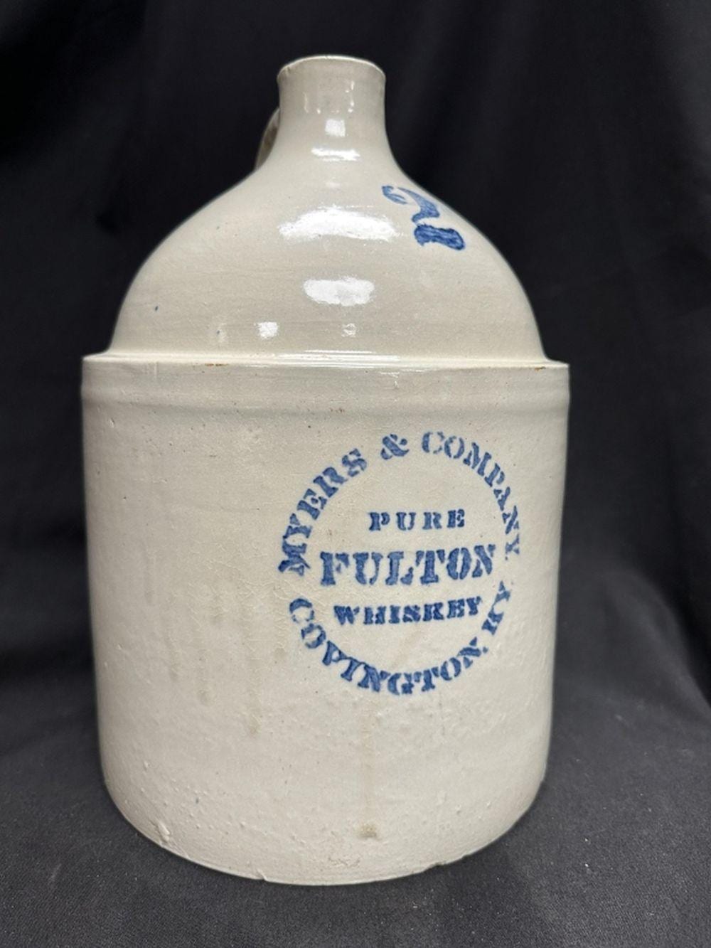 White ceramic advertising whiskey jug with a blue printed label and decorative blue motif on the neck, against a dark background.