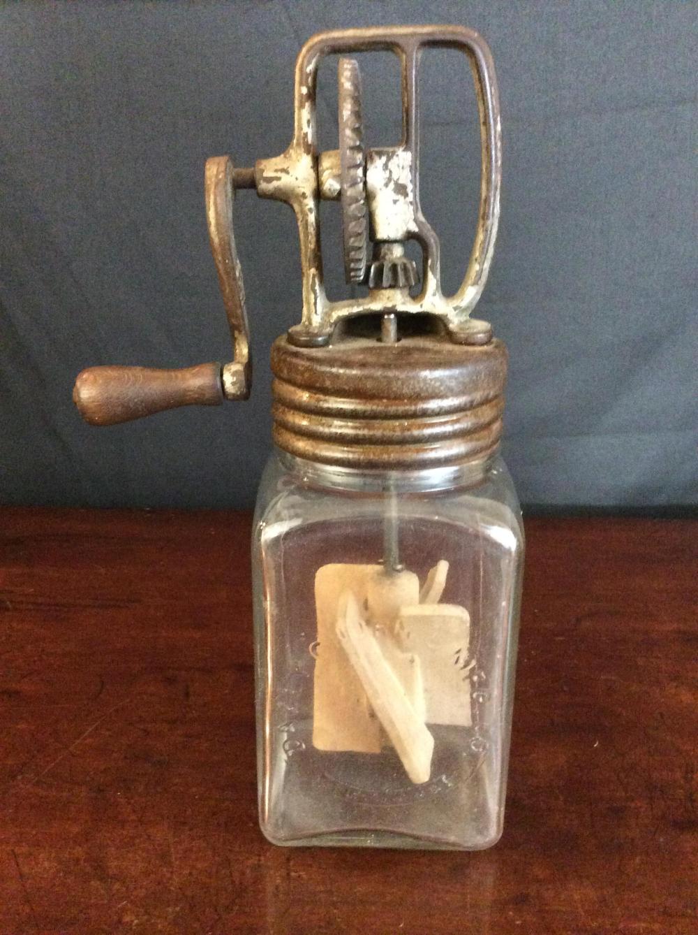 Antique metal hand butter churn with a glass jar and wooden handle, photographed against a gray background.