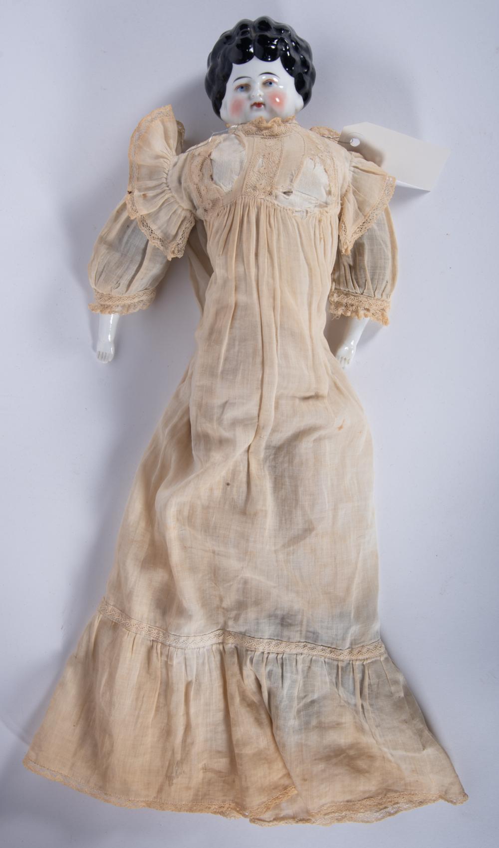 Porcelain head doll with curly dark hair and a pale face, dressed in a light cream lace-trimmed gown with long sleeves, standing against a plain background.