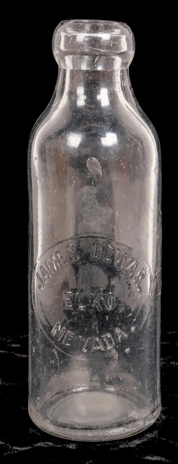 Clear glass soda bottle with embossed lettering on the front and a rounded shoulder, shown against a dark background.
