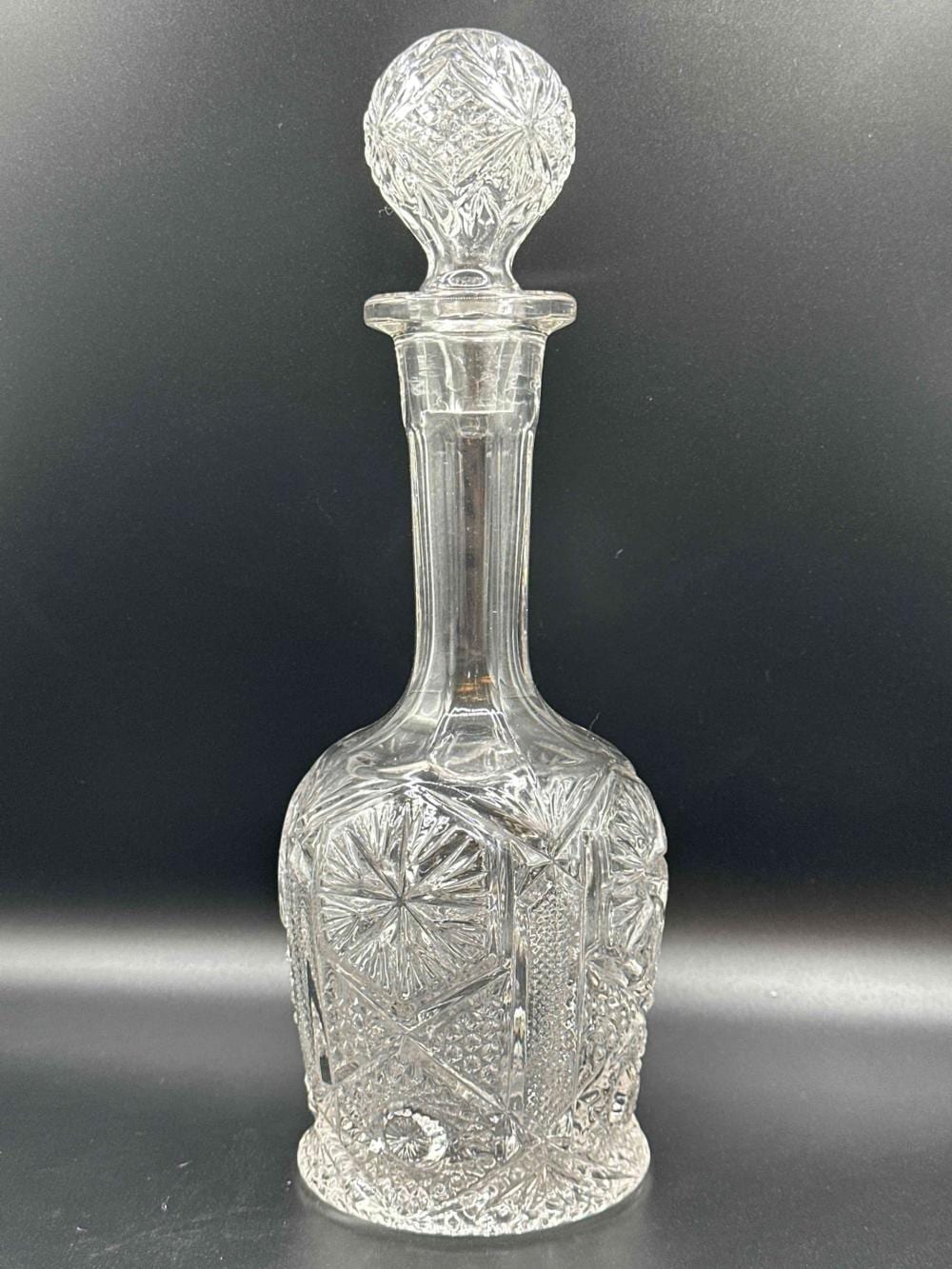 Clear pressed-glass decanter with faceted texture and a rounded stopper, shown on a dark background.