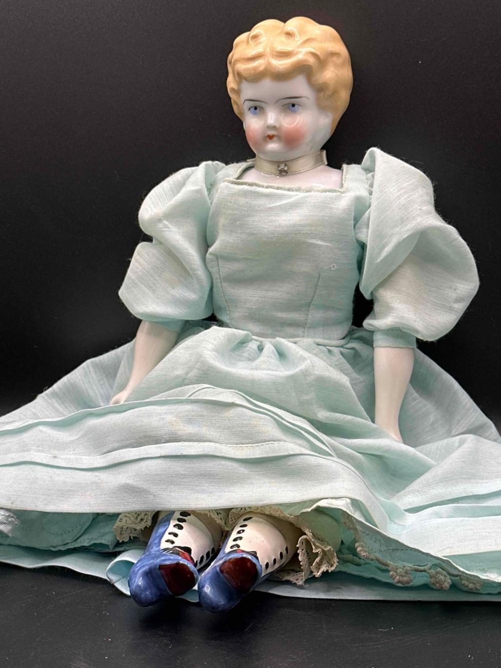 Porcelain doll with painted face and blonde hair wearing a light blue dress and lace cuffs, seated with two porcelain shoes at its feet.