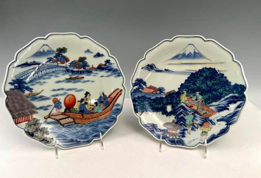 Pair of scalloped porcelain dishes with blue-and-white landscape scenes and boats, mounted on small metal stands.