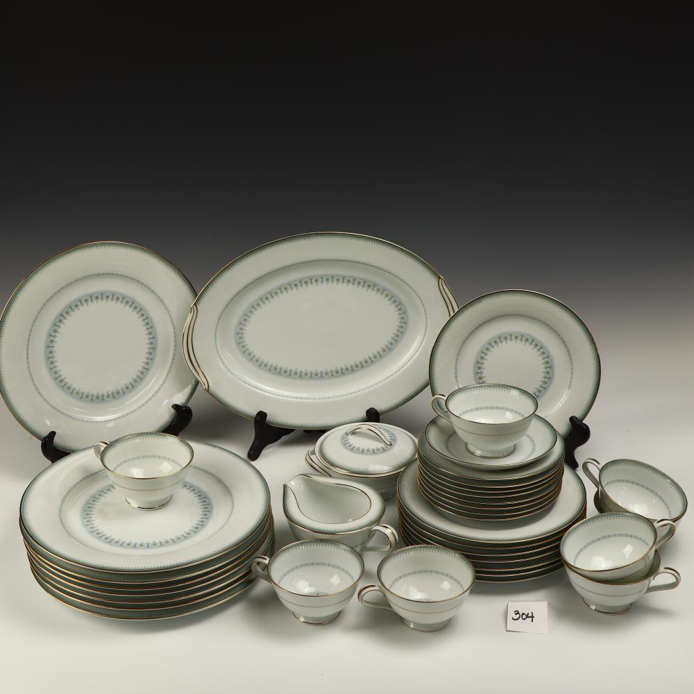 Set of white ceramic dinnerware with oval blue border pattern and gold trim, including plates, bowls, and cups arranged on a display.