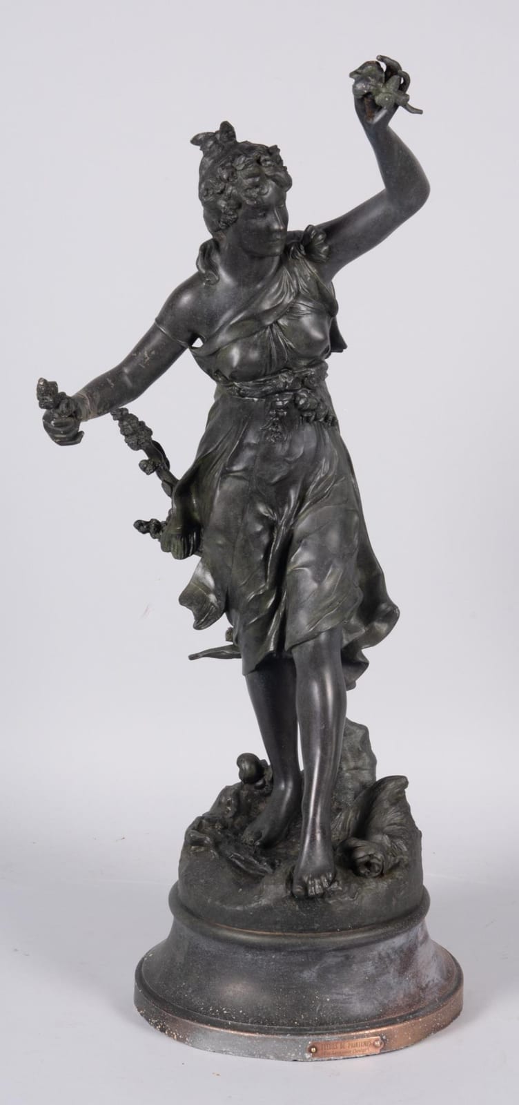 Bronze-colored figure of a dancing woman with curled hair, arms raised holding a small bouquet of flowers, standing on an oval base.