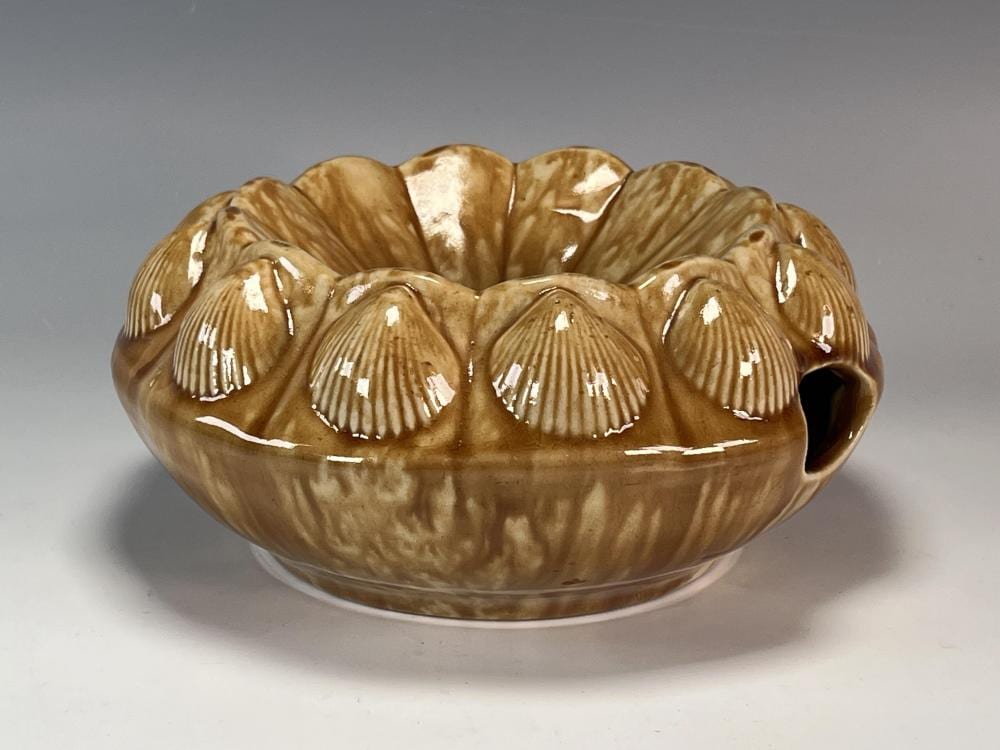 Glazed brown stoneware pottery bowl or spittoon with scalloped seashell-like ridges and a small side opening on gray background.