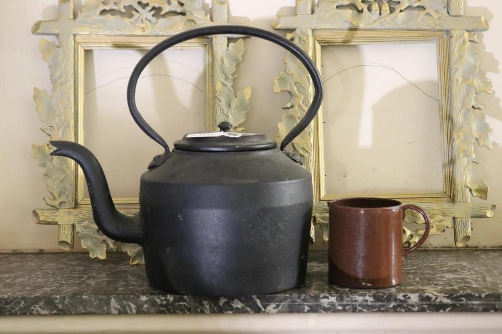 Black cast iron kettle with curved handle and spout, positioned in front of a decorative mirror; a brown enamel jug sits beside it.