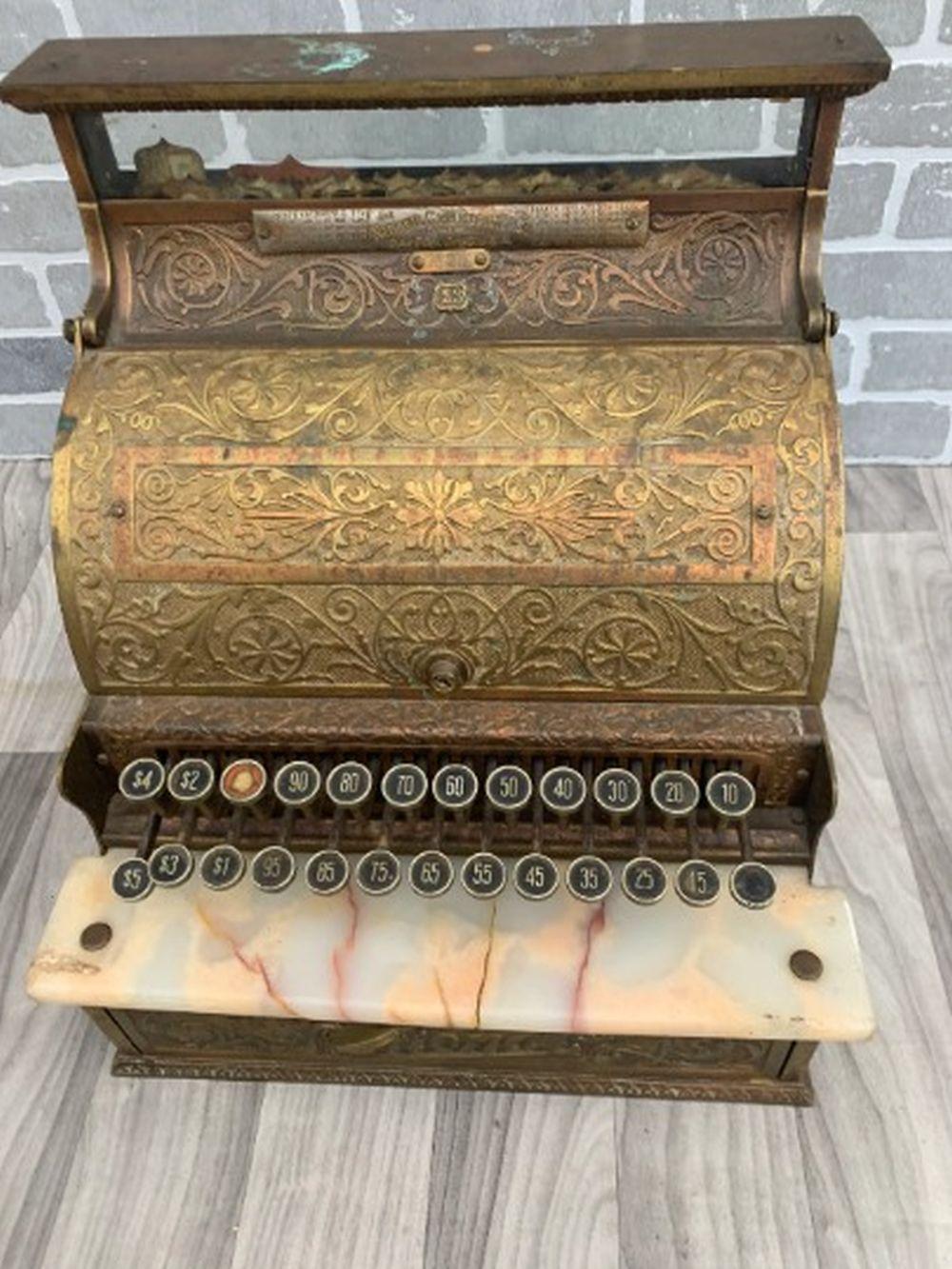 Antique-style cash register with ornate gold-toned panels, hinged drawer, and rows of numbered keys and knobs.