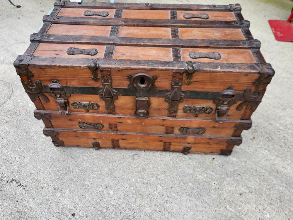 Antique wooden steamer trunk with metal straps, handles, and latch hardware, shown on a gray floor.