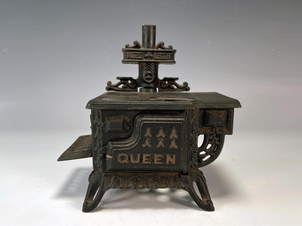 Vintage cast iron miniature toy stove with a chimney pipe and “QUEEN” panel, shown on four legs.